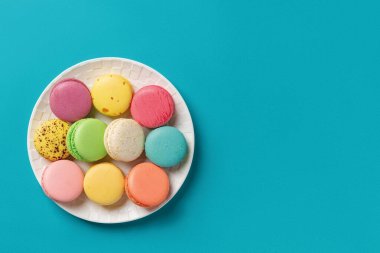 Parlak turkuaz arka planda beyaz yuvarlak bir tabakta renkli badem kurabiyeleri. Çok renkli badem kurabiyeli tebrik kartının bir kopyası. Lezzetli Fransız bademli kurabiyeleri. Uzayı kopyala Üst görünüm.