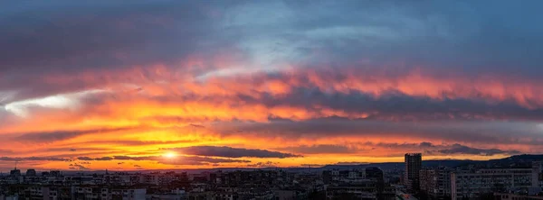 Dramatic sunset fire in the sky over a small city. Evening sun is low on the horizon. Panoramic landscape with urban skyline. Vivid red orange and dark blue sky during sundown. Scenic skyscape.