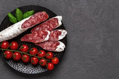 Sliced thin catalan fuet sausage and bunch of red cherry tomatoes on a black plate over dark rough surface. Traditional spanish dry-cured pork salami with white noble mold. Tasty meat delicacy. Copy space. Top view. clipart