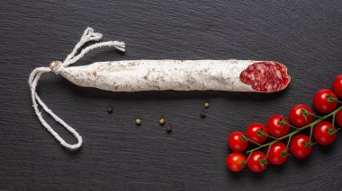Siyah bir sayfada ince katalanlı sosis ve bir demet kırmızı kiraz domatesleri. Beyaz asil kalıplı geleneksel İspanyol kurutulmuş domuz salamı. Lezzetli et lezzeti. Üst görünüm. 