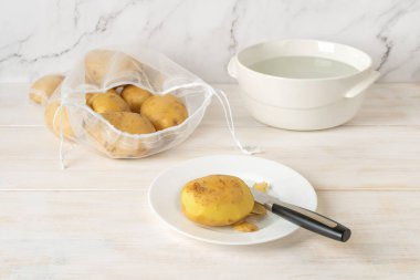 Yarısı soyulmuş çiğ patates yumrusu ve tabakta bıçak ve yeniden kullanılabilir naylon poşet içinde soyulmamış organik patatesler beyaz ahşap mutfak masasının üzerinde. Sıfır atık ve sürdürülebilir yaşam tarzı kavramları. Ön görünüm.