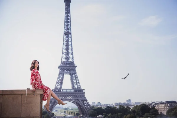赤いドレスを着た黒い髪の女性がパリの背景にエッフェル塔に座っています — ストック写真