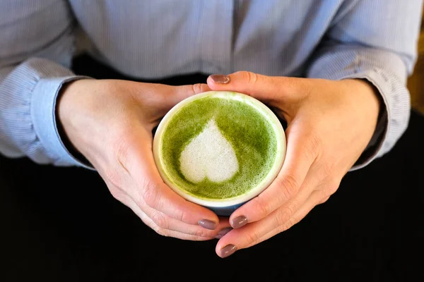 Mujer sosteniendo taza azul con latte verde de moda caliente con corazón de arte en la espuma sobre fondo de mesa de madera negro, bebida saludable con arte latte —  Fotos de Stock