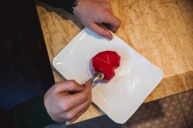 Masanın üzerinde kalp şeklinde kırmızı bir pasta var. Kaşıklı insan elleri turtaya uzanıyor. Sevgililer Günü