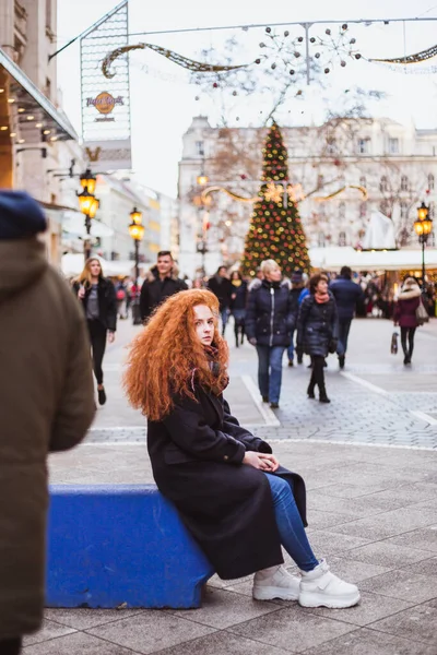 Rött hår flicka njuter av den europeiska julmarknaden. suddiga ljus på julgran på bakgrunden — Stockfoto