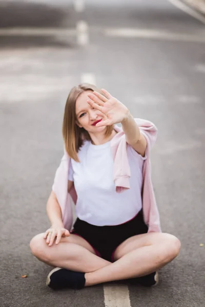 Joven mujer feliz en camiseta blanca se sienta en el medio de la carretera de asfalto y muestra parar con su mano —  Fotos de Stock