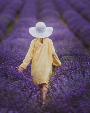 Sarı elbiseli gülümseyen küçük kız ve lavanta tarlasında koşan beyaz şapka