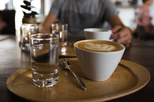 Filiżanka kawy cappuccino i szklanka wody na drewnianej tacy. Mężczyzna trzymający filiżankę kawy na tle — Zdjęcie stockowe