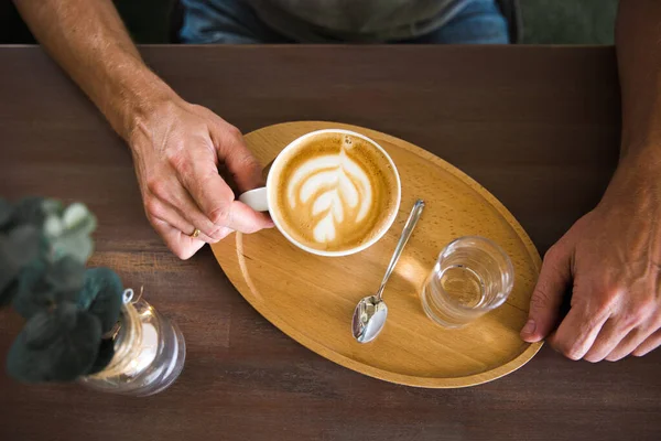 Mężczyzna trzymający filiżankę kawy cappuccino i szklankę wody na drewnianej tacy. Widok z góry — Zdjęcie stockowe