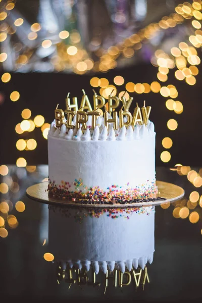 Birthday cake with candles, garland with bright bokeh lights on the background. The white cake is decorated with colored sprinkles and stands on a reflective surface. — Stock Photo, Image