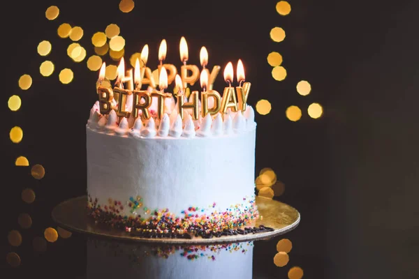Birthday cake with candles, garland with bright bokeh lights on the background. The white cake is decorated with colored sprinkles and stands on a reflective surface.