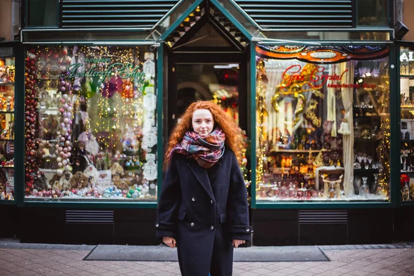 Chica pelirroja disfrutando del Mercado Europeo de Navidad. Luces borrosas y juguetes de colores en la tienda de Navidad en el fondo —  Fotos de Stock