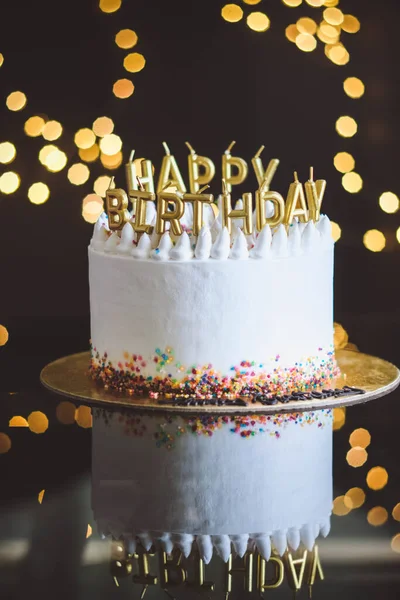 Tort urodzinowy ze świecami, girlanda z jasnymi światłami bokeh na tle. Białe ciasto jest ozdobione kolorowymi posypkami i stoi na powierzchni odblaskowej. — Zdjęcie stockowe