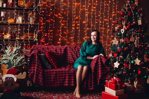 Mujer sonriente en elegante vestido verde en el sofá en el árbol de Navidad. —  Fotos de Stock