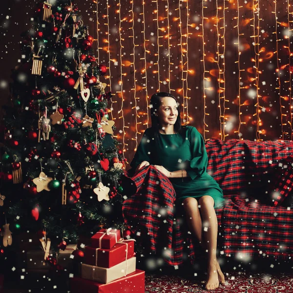 Leende kvinna i elegant grön klänning på soffa vid julgran. — Stockfoto