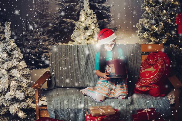 Щаслива маленька дівчинка в капелюсі Санта Клауса з присутнім на дивані різдвяні подарунки проти яскравого освітлювального садівника.. — стокове фото