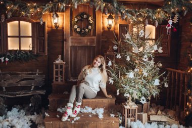 Genç ve güzel bir kız, yeni yıl için dekore edilmiş bir köy evinin bahçesinde oturuyor..