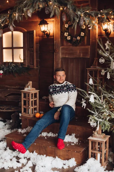 Um jovem bonito em uma roupa quente senta-se nos degraus no pátio da casa guirlanda antes do Ano Novo — Fotografia de Stock