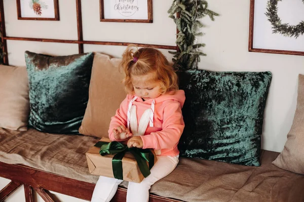 Pequeña chica bonita sith pelo ondulado se sienta del sofá abre una caja con un regalo — Foto de Stock