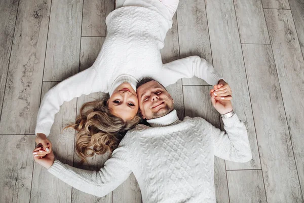 Pareja sonriente en suéteres en el suelo. Feliz pareja amorosa en suéteres de invierno de punto blanco tirados en el suelo tomados de la mano. — Foto de Stock