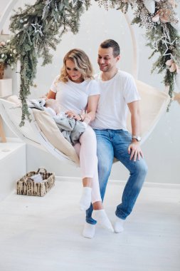 Bebekli neşeli ebeveynler Noel salıncağında. Çekici bir ailenin portresi, köknar dallarıyla süslenmiş rahat salıncakta oturan bebekle eğleniyor..