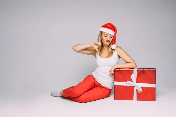 Noel şapkalı komik kadın büyük kırmızı bir kutuyla oturur ve bundan hoşlanmaz. Beyaz arka planda izole edilmiş bir resim. — Stok fotoğraf
