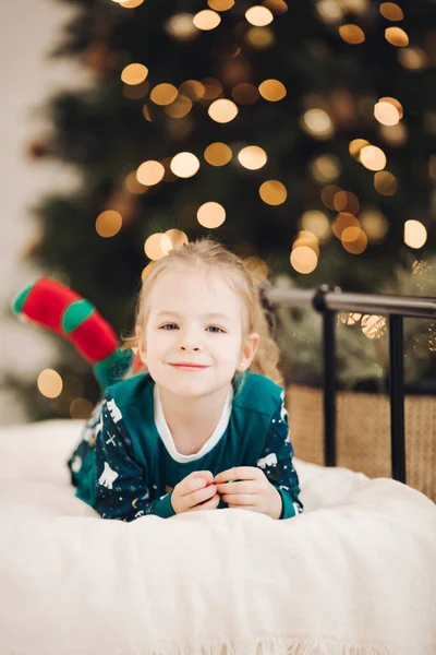 Klein blank meisje ligt op het bed in de buurt van de kerstboom en glimlacht — Stockfoto