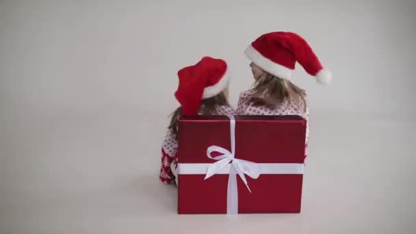 Twee vrolijke meisjes zitten achter een grote rode doos met een nieuwjaarscadeau en zwaaien met hun hoofd — Stockvideo