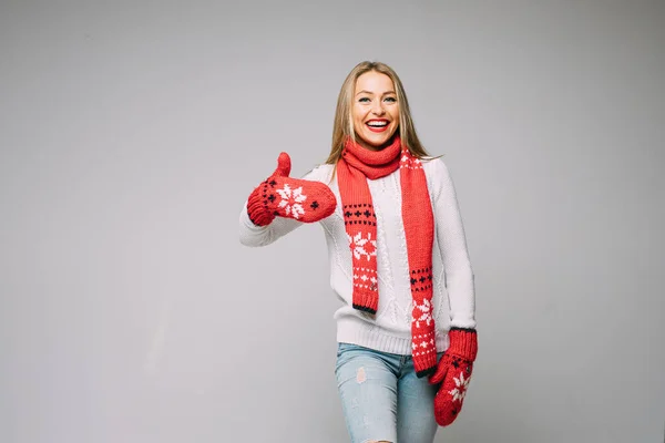 Atractiva mujer en suéter blanco, bufanda roja y mitones rojos con diseño de Navidad muestra como — Foto de Stock