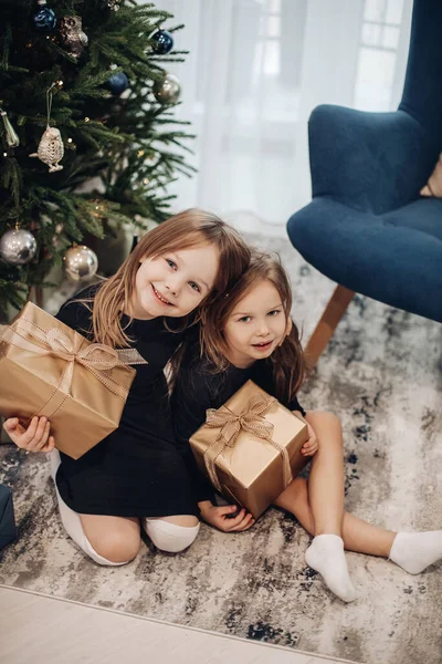 Маленькі кавказькі дівчата дивляться свої подарунки біля ялинки вдома і посміхаються разом — стокове фото