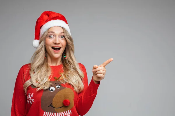 Menina loira no chapéu de Santa mostrando espaço em branco com o dedo. olhando para a câmera com emoção e boca aberta. — Fotografia de Stock
