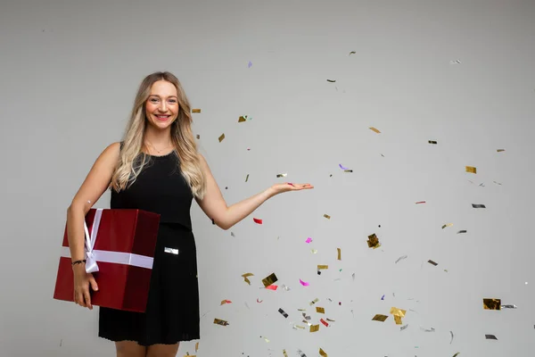 Radosna kobieta w czarnej sukience raduje się z pudełka ze swoim prezentem świątecznym z dużą ilością konfetti wokół niej — Zdjęcie stockowe