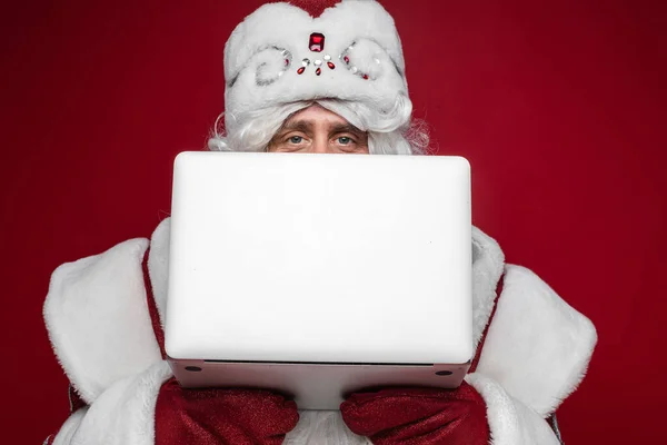 Santa Claus hiding behind laptop with copy space on red studio background, peek out checking xmas email accepting orders — Stock Photo, Image