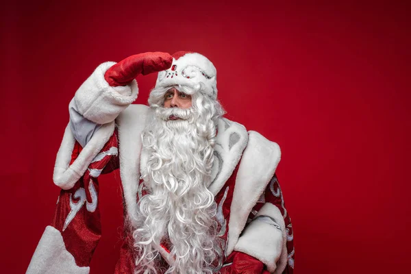 Noel Baba, elini alnına koyup uzaklığa bak. Kırmızı arka planda. Tatil reklamları için yer ayır. — Stok fotoğraf