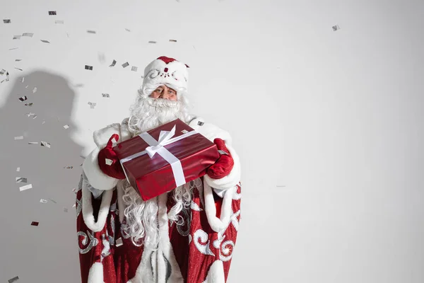 Ojciec Frost w długim ciepłym płaszczu, czerwone rękawiczki i kapelusz posiada prezent świąteczny z dużą ilością konfetti wokół niego — Zdjęcie stockowe