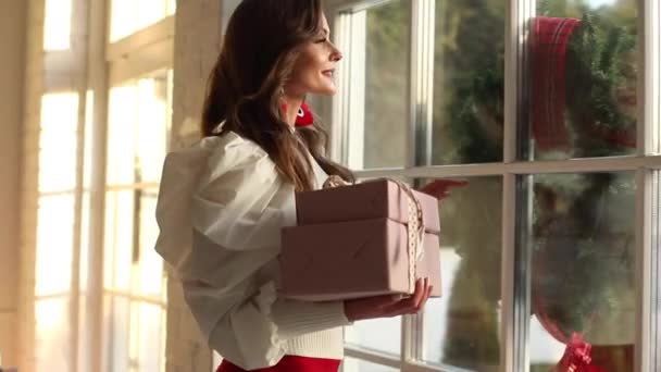 Álomszép, fiatal nő, ajándékokkal a kezében, mosolyog és bámul az ablakon, csodára várva. — Stock videók