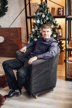 Kendinden emin erkek, Noel ağacının arka planında rahat bir koltukta oturuyor.