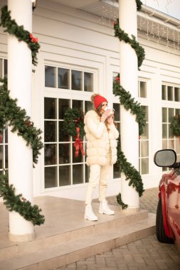 Beyaz kürklü ve kırmızı şapkalı çekici kadın dışarıda gülümsüyor.