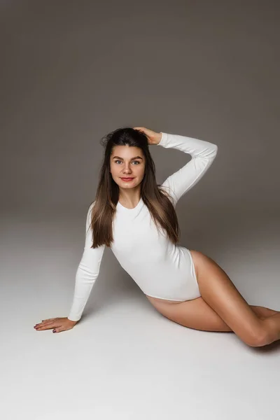 Mooi jong fit meisje in wit bodysuit, gebruind slank vrouwelijk model met lange gezonde haren poseren op studio vloer — Stockfoto