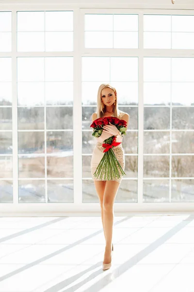 Bella ragazza bionda modello di moda femminile che indossa abito corto tenere bouquet di rose festive posa vicino a una grande finestra — Foto Stock