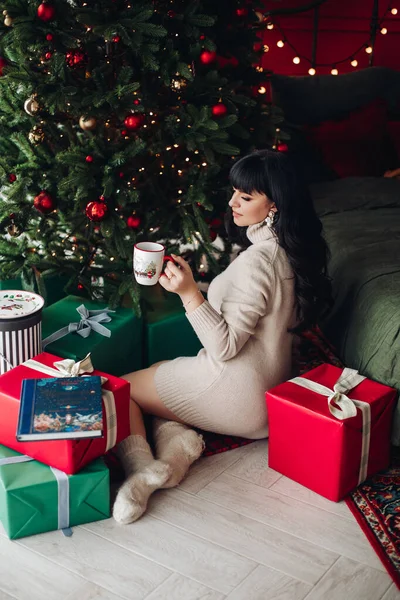 Elinde kahve fincanıyla Noel ağacının yanında oturan bir kadın. — Stok fotoğraf