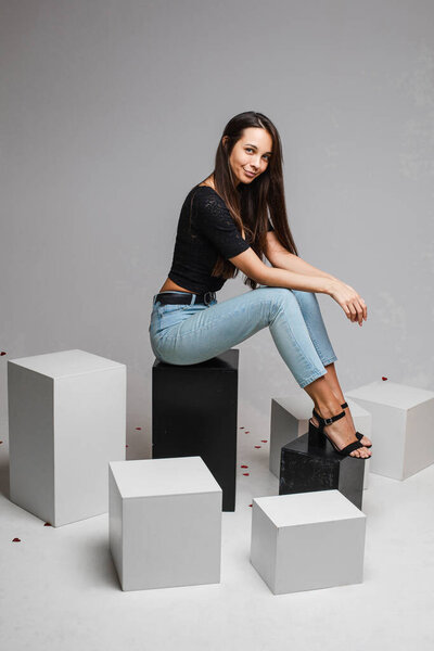 Smiling young fit girl with long hairs posing sitting on black and white cubes on gray studio background with copy space