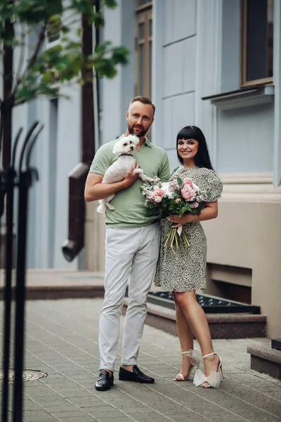 Glad kärleksfull romantisk par promenader med husdjur liten hund utomhus. Flicka med blommor bukett och kille med valp på dejting — Stockfoto