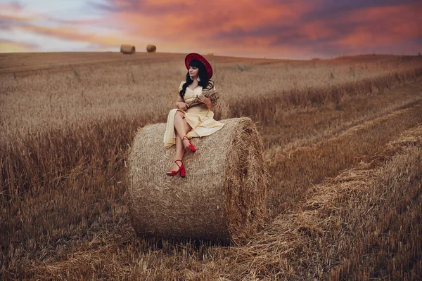 Krásná mladá žena sedí na velké kupce sena na poli — Stock fotografie