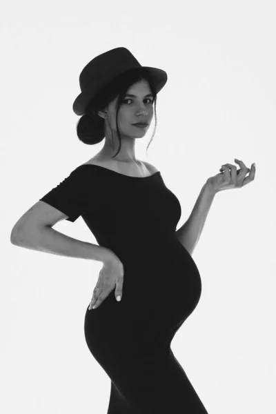 Frumoasă fată gravidă tânără cu burtă purtând pălărie și rochie neagră așteptând un copil. Sarcina elegantă. Negru și alb — Fotografie, imagine de stoc