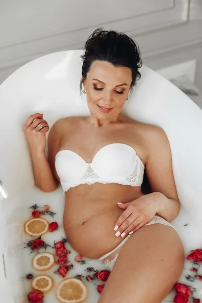 Mulher grávida atraente relaxante no banho com água branca e um monte de laranjas e flores — Fotografia de Stock