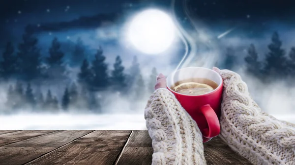 Red cup with coffee, tea in female hands on a snowy background, winter forest. A red cup in hands against the background of a winter forest landscape on soft, knitted sleeves. Coffee, tea on a wooden