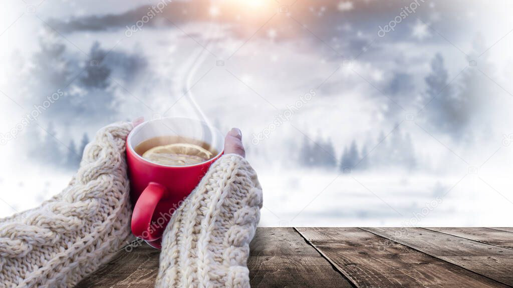 Red cup with coffee, tea in female hands on a snowy background, winter forest. A red cup in hands against the background of a winter forest landscape on soft, knitted sleeves. Coffee, tea on a wooden 