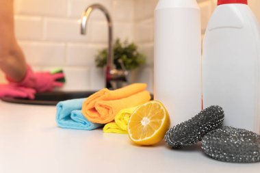 Mutfak tezgahında temizlik ürünleri, sitron ve süngerler odak ve arka planda bulanık kadın elleri mutfağı temizliyor..