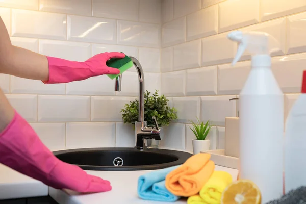 Womens hands cleaning kitchen tap on a black sink with rubber gloves and in front blurred cleaning products for disinfection.